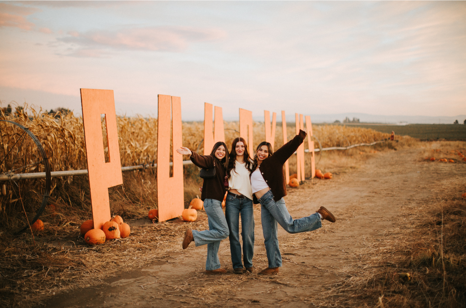 Three people posing in a pumpkin patch with pink wooden cutouts in the background.